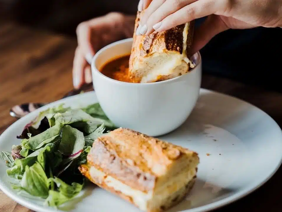 A person dipping a grilled cheese from Culture Meat & Cheese into a bowl of tomato soup