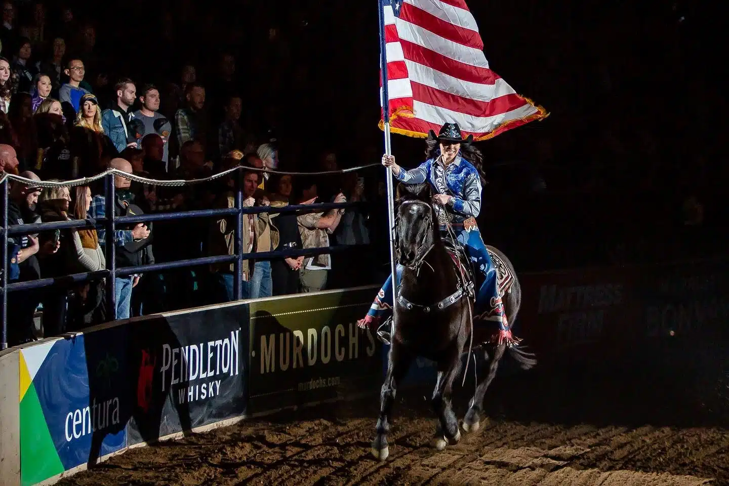 A woman on horseback carrying an American flag and riding past a crowd at the National Western Complex