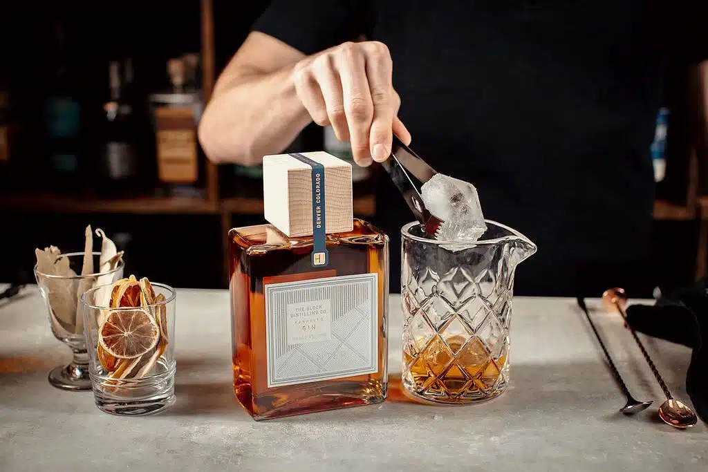 A bartender places an ice cube into a glass, which rests next to a bottle of gin by The Block Distilling Co.