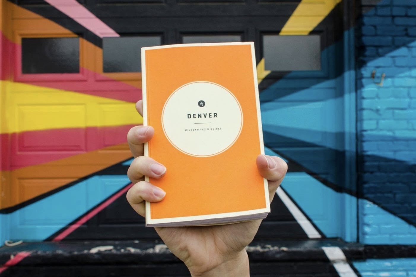 A person holding up a Wildsam Denver field guide against a vibrant mural